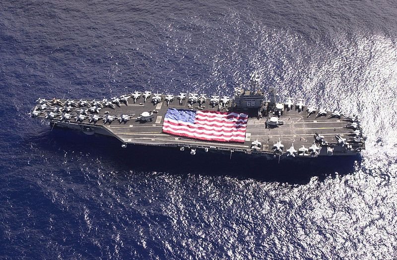 An aircraft carrier under way with the national ensign spread across the flight deck